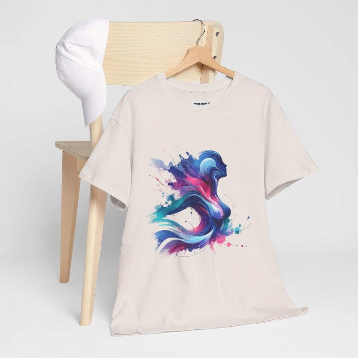 Ice grey cotton t-shirt with colorful abstract graphic print draped on wooden chair next to a white baseball cap