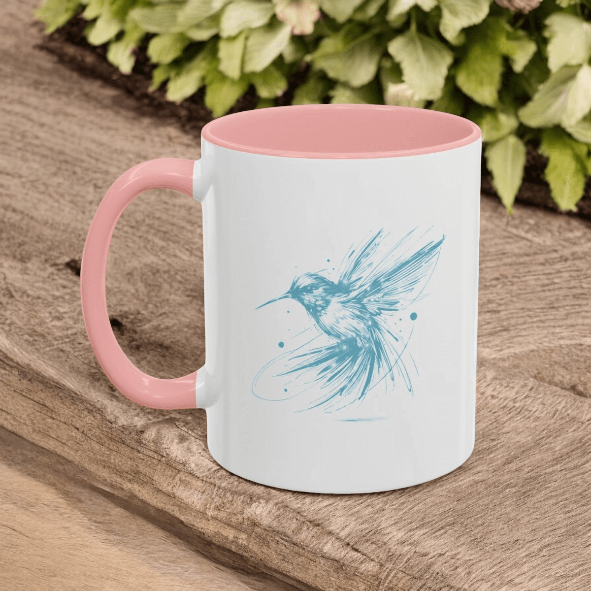 White ceramic mug with pink interior and handle, features blue hummingbird sketch, placed on wood with greenery background
