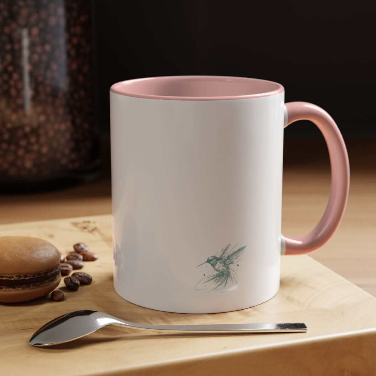 White ceramic mug with pink handle and hummingbird graphic placed on wooden surface with brown macaron and spoon