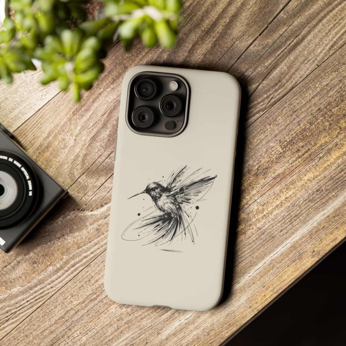 Beige phone case for iPhone featuring a printed hummingbird design, shown on wooden table near camera and plant