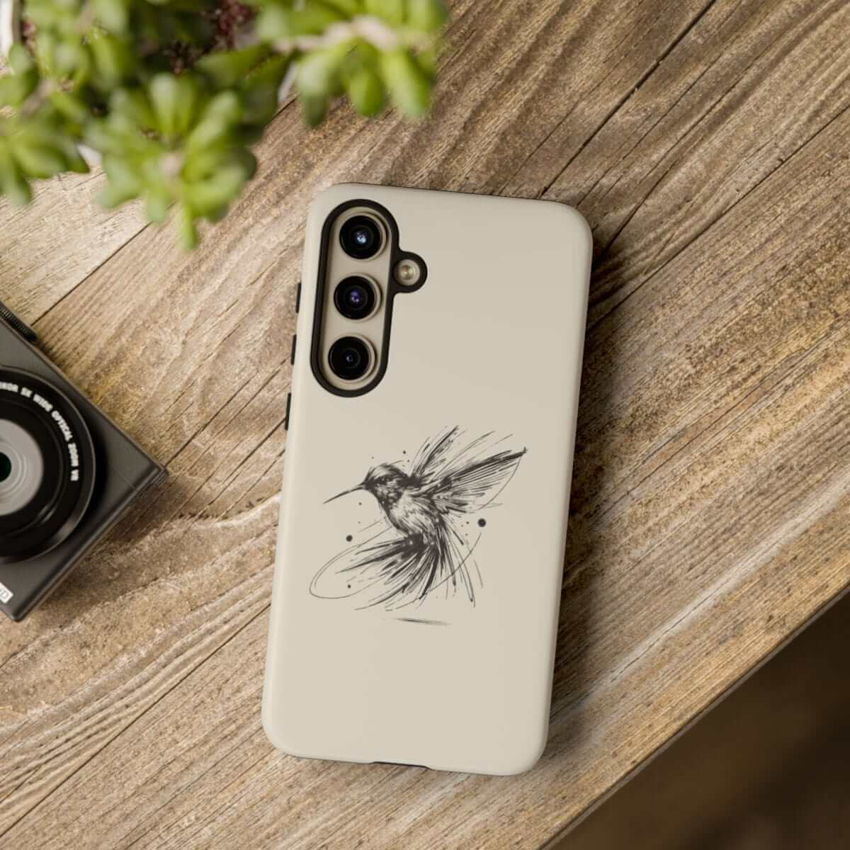 Beige phone case made of matte silicone featuring a minimalist hummingbird sketch placed on wooden surface near plant