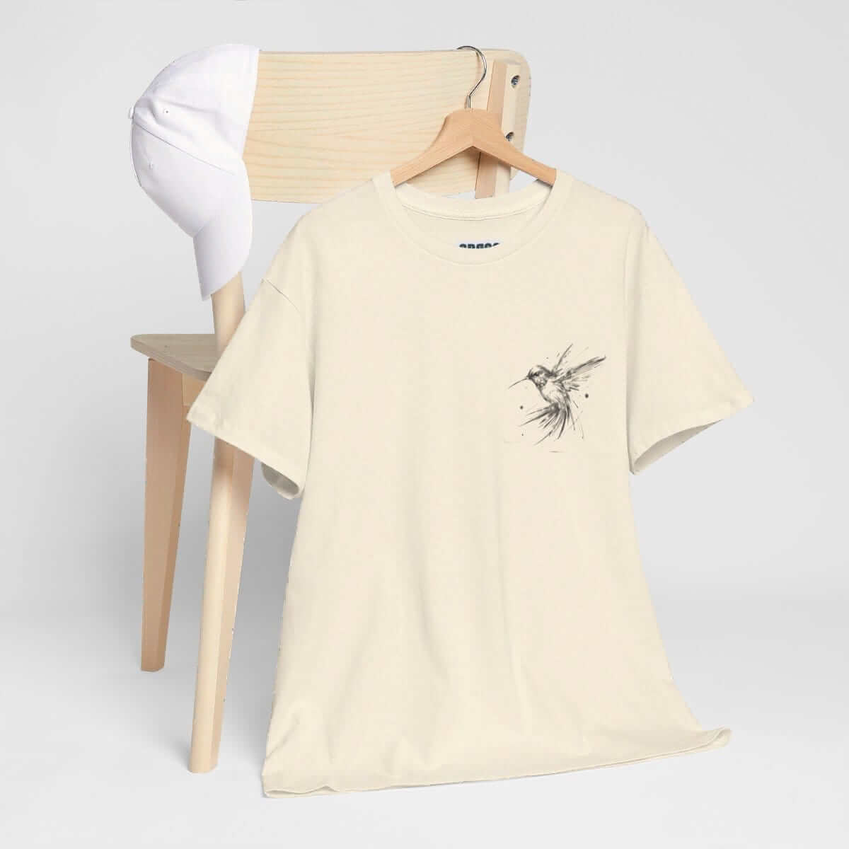 Beige cotton T-shirt with minimalist bird graphic draped on a chair beside a white baseball cap