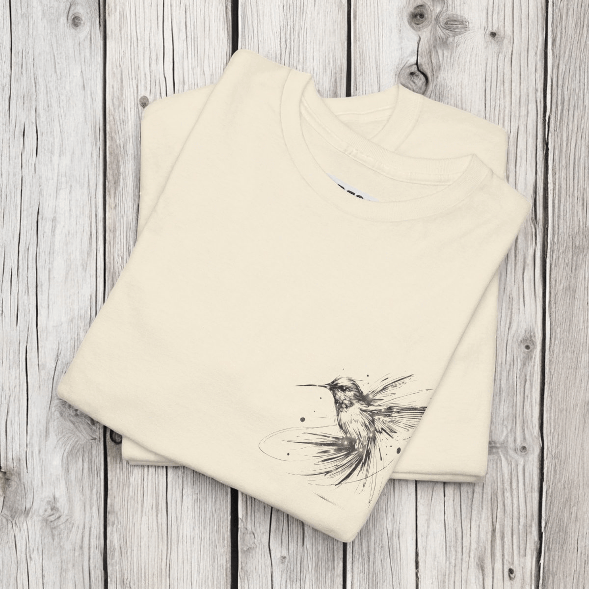 Cream cotton unisex t-shirt featuring a black line art hummingbird sketch on the chest, laid flat on wood surface