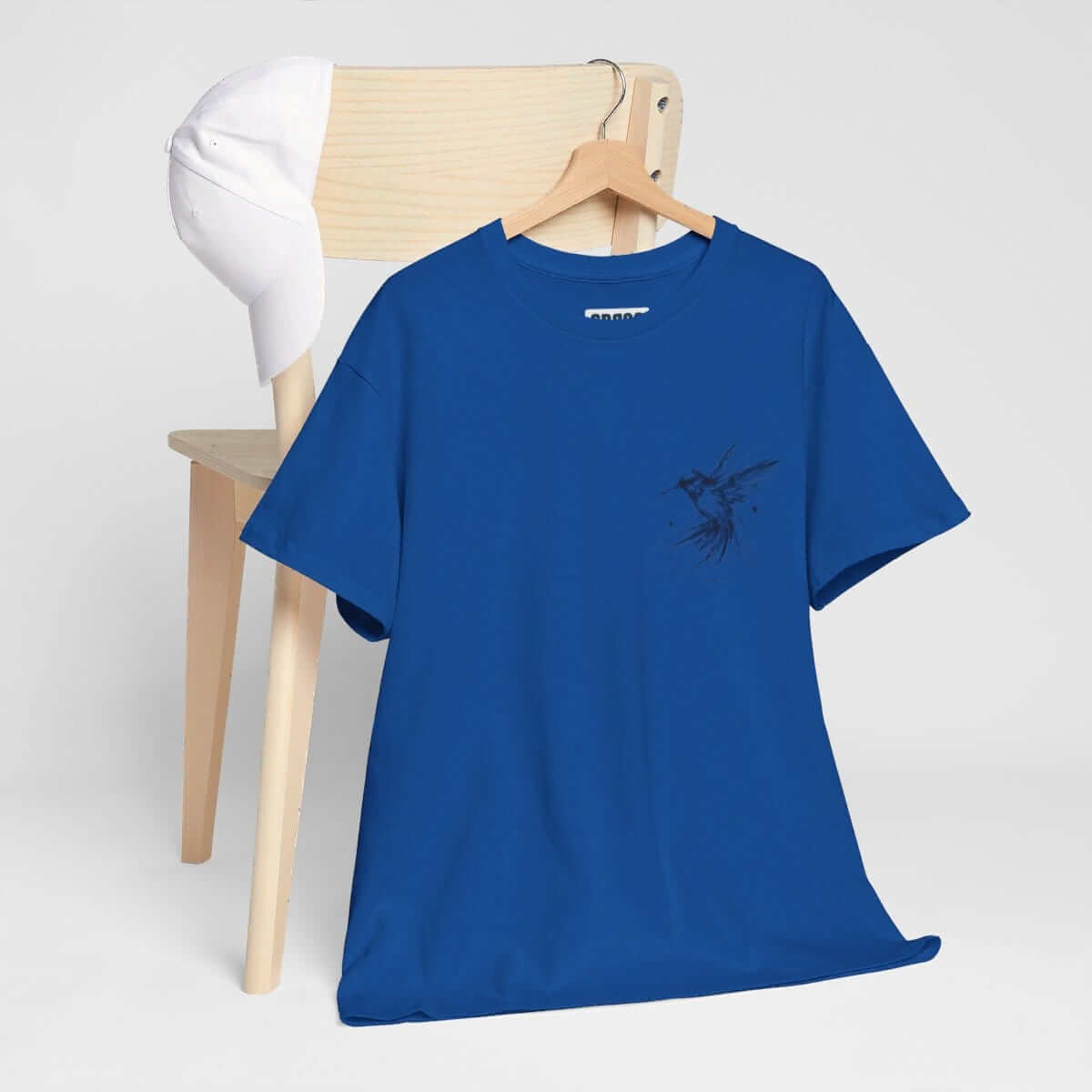 Blue cotton t-shirt with short sleeves and illustrated bird graphic draped over a wooden chair next to a white cap