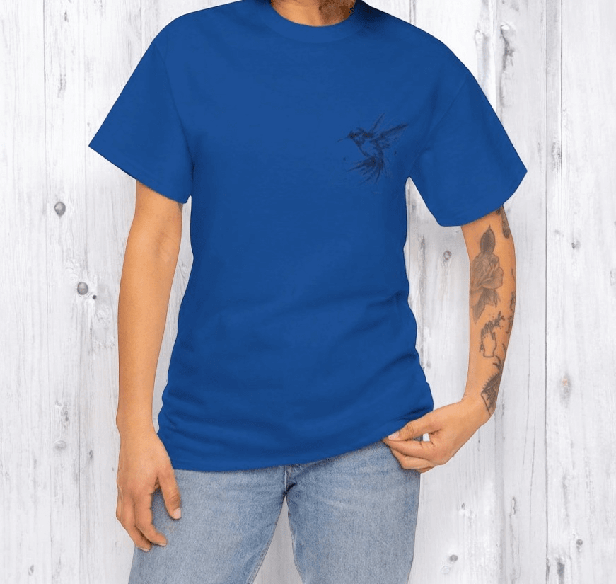 Blue cotton t-shirt with short sleeves and a small dark chest graphic worn by tattooed person in outdoor setting