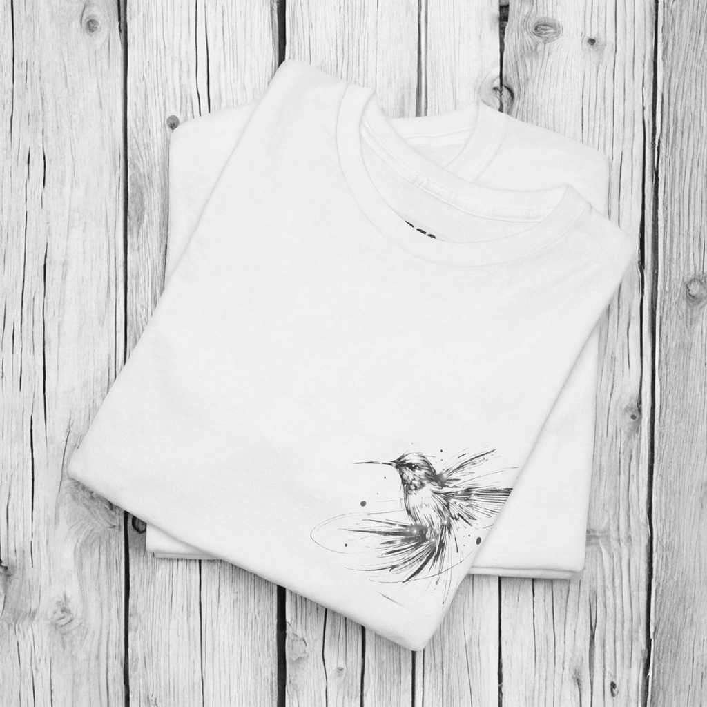 White cotton T-shirt with black hummingbird sketch printed on front, laid flat on a rustic wooden surface