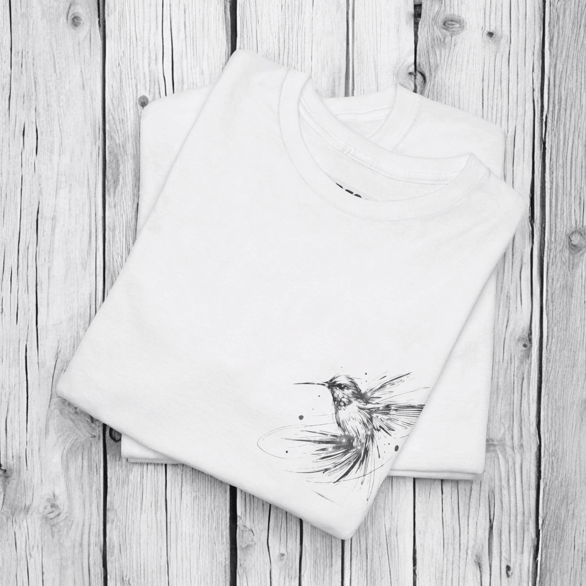White cotton T-shirt with black hummingbird sketch printed on front, laid flat on a rustic wooden surface