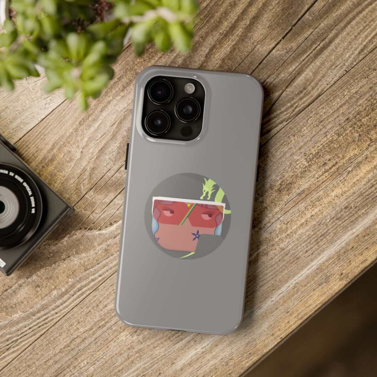 Gray smartphone with a matte finish and colorful triangle sticker, resting on wooden surface beside camera and plant