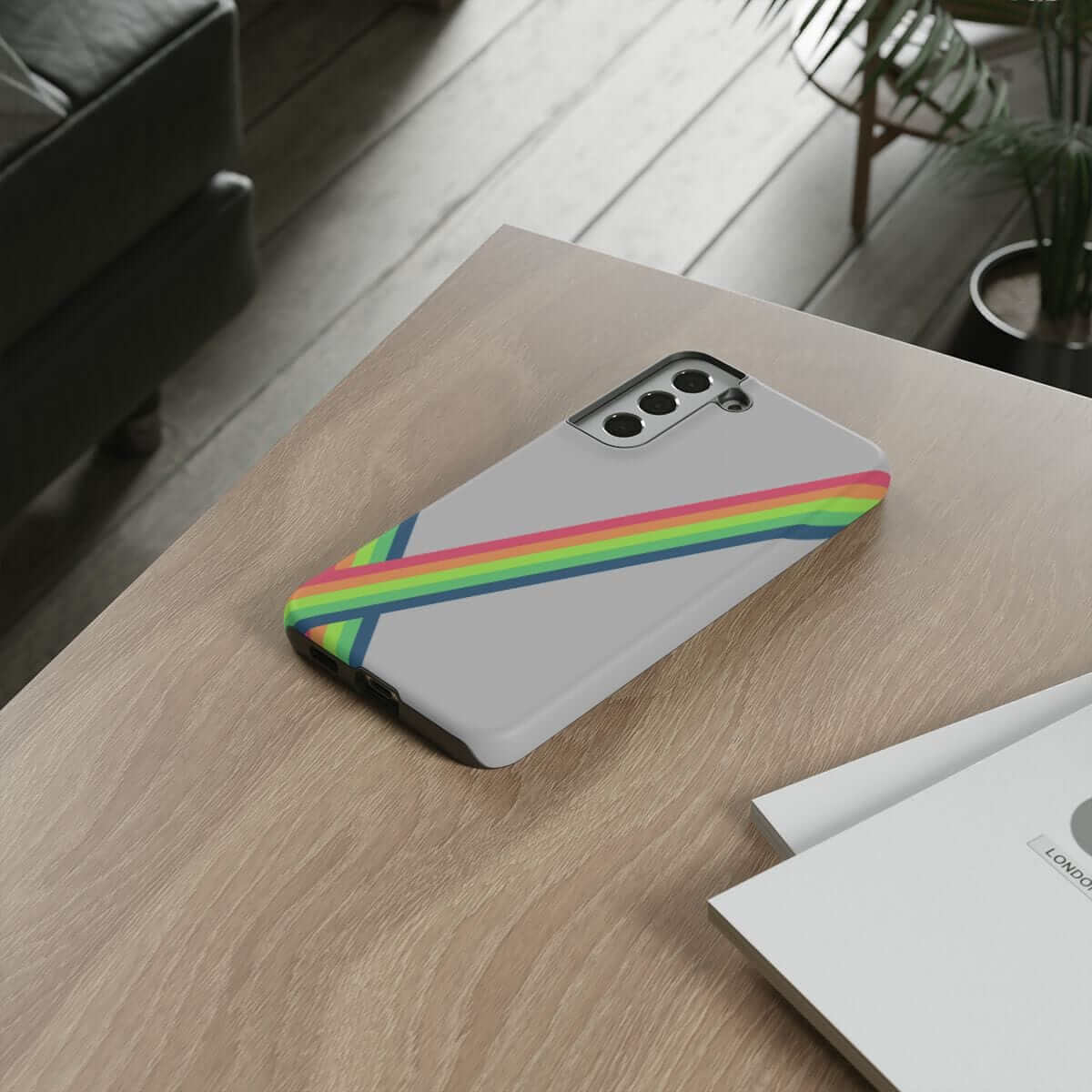 Grey phone case made of matte plastic with vertical rainbow stripes, displayed on wooden table near plant and papers