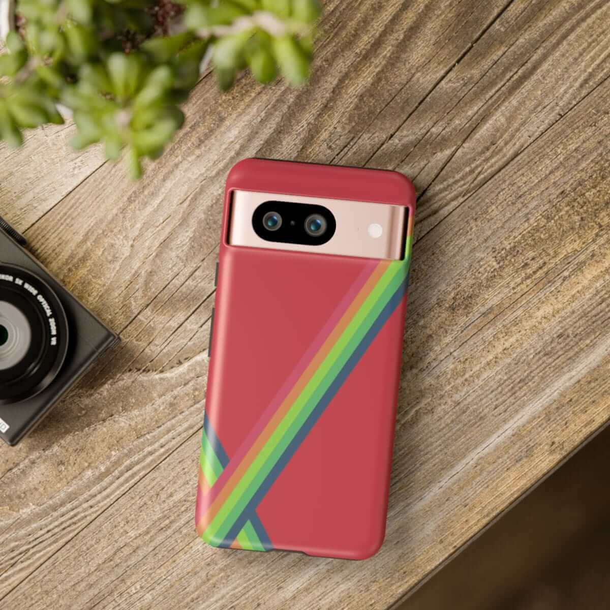 Phone case in red with green and yellow diagonal stripes, hard plastic design, shown on wooden surface beside a plant