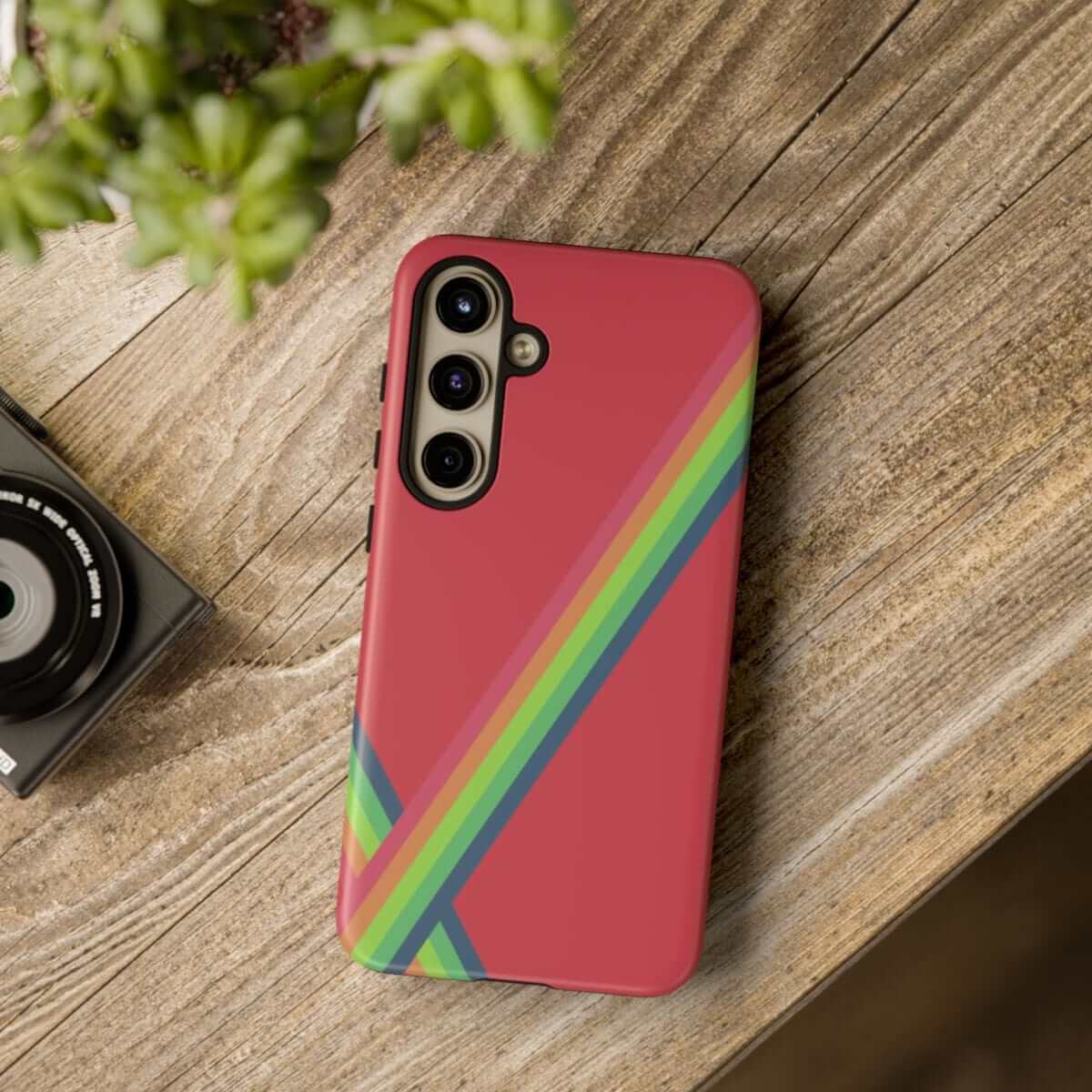 Smartphone with red protective case featuring diagonal rainbow stripes, shown on a wooden table near a camera and plant